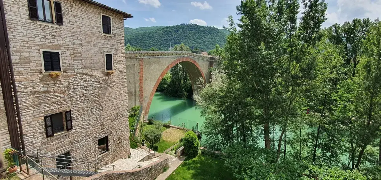 Un antico ponte ad arco domina un fiume verde smeraldo, perfetto per il trekking nelle Marche. Un borgo in pietra completa la scena.
