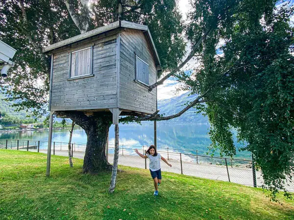 Bambina corre verso una casa sull'albero in legno, con un lago e montagne sullo sfondo.