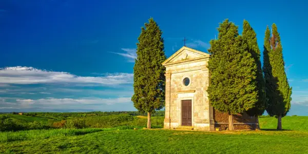 Cappella della Madonna di Vitaleta, un gioiello a San Quirico d'Orcia, circondata da cipressi e colline verdi sotto un cielo azzurro.