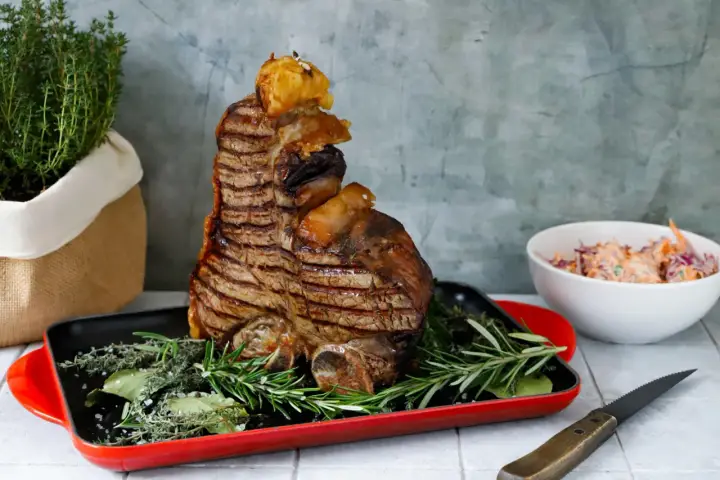 Una succulenta bistecca fiorentina, grigliata alla perfezione, adagiata su erbe aromatiche in una teglia rossa. Accanto, un'insalata fresca e un coltello.