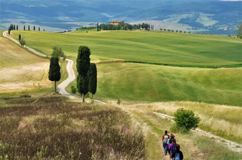 Un gruppo di escursionisti vive le **esperienze Val d'Orcia** su un sentiero tra colline verdi e cipressi, con una casa colonica in lontananza.